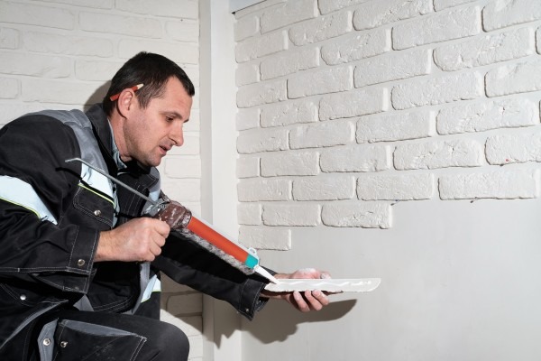 Contractor applying sealant to a white brick wall using a caulking gun indoors